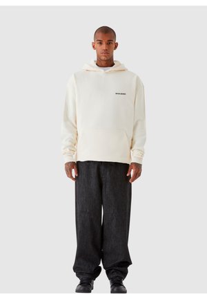 Cremefarvet hoodie med frontlomme, med et lille sort logo. Båret med mørke, løstsiddende denimbukser og sort fodtøj.