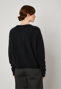 Melna, pūkaina džemperis ar apaļu kakla izgriezumu un garām piedurknēm, redzams no aizmugures. Valkāts ar tumši zaļiem biksēm; redzama mīksta tekstūra.