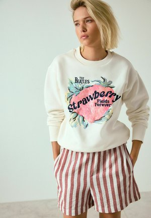 Young woman wearing cream sweatshirt with "The Beatles Strawberry Fields Forever" print and red and white vertical striped shorts, hands in pockets.