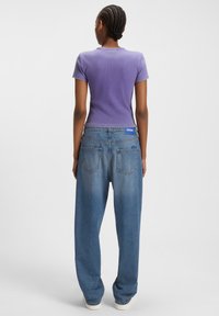 Femme avec des cheveux tressés portant un t-shirt violet ajusté à manches courtes et un jean bleu décontracté avec une étiquette HUGO bleue, se tenant de dos.