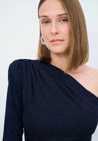 Vestido azul marino de un solo hombro con tejido texturizado y brillante. Presenta detalles fruncidos en el hombro y una silueta ajustada.