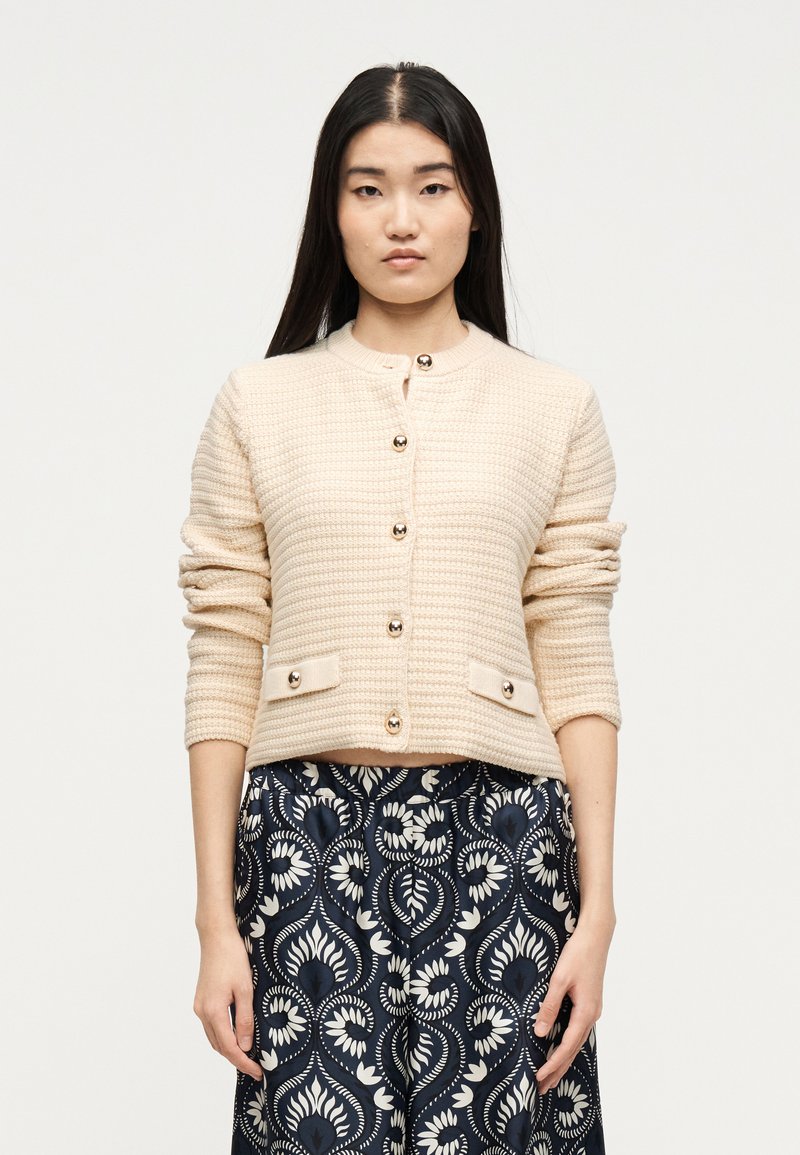 Γυναίκα με μακριά σκούρα μαλλιά, ντυμένη με μπεζ υφασμάτινο ζακέτα με κουμπιά και μπλε σκούρο παντελόνι με λευκό ανθισμένο σχέδιο.