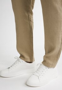 Baskets blanches en cuir avec une surface texturée, lacets plats et une semelle en caoutchouc blanche épaisse, associées à un pantalon en lin marron clair.