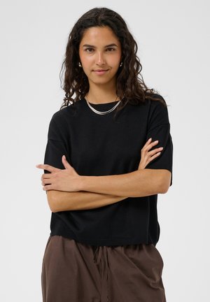 Femme aux longs cheveux bouclés portant un haut noir à manches courtes et un pantalon marron, bras croisés, sur un fond clair uni.