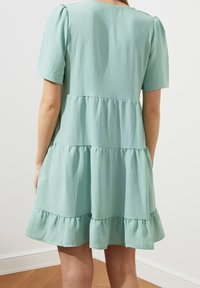 Světle modré šaty s vrstveným designem, vyrobené z měkké tkaniny; mají krátké nabírané rukávy a volánkový lem, s uvolněným střihem a hladkou texturou.
