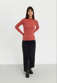 Maglione in corallo a costine con maniche lunghe, abbinato a una gonna lunga nera con spacco laterale, indossato con stivaletti neri alla caviglia. Sfondo semplice.