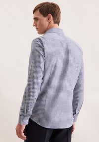 Uomo rivolto a sinistra, che indossa una camicia a maniche lunghe a scacchi blu e bianchi e pantaloni neri, su uno sfondo chiaro e semplice.