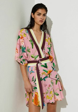 Kobieta w różowej kwiecistej sukience kopertowej z głębokim dekoltem w kształcie litery V i bordowymi oraz żółtymi obszyciami, stojąca przy białej ścianie.