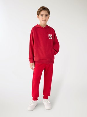 Ragazzo in piedi con una mano in tasca, che indossa una felpa con cappuccio rossa con il numero "99" e pantaloni rossi abbinati e scarpe da ginnastica bianche.