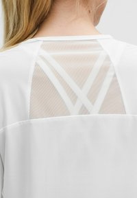 Bijela majica s prozirnim mrežastim leđima i ukrštenim bijelim trakama. Tkanina izgleda glatko s opuštenim dizajnom.