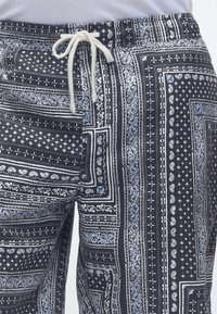 Pantalon bleu marine avec des motifs paisley blancs et géométriques, avec un cordon blanc noué en nœud à la taille.