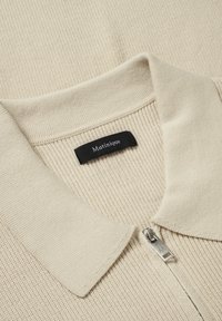 Bēša ribota polo krekls ar īsu rāvējslēdzēju un mīkstu apkakli. Satur iekšpusē piešūtu melnu etiķeti ar zīmola nosaukumu "Matinique".