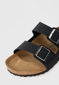 Sandale en cuir noir à deux sangles avec boucles métalliques réglables, semelle en liège et semelle extérieure en caoutchouc texturé. Présente un design plat et une forme arrondie.