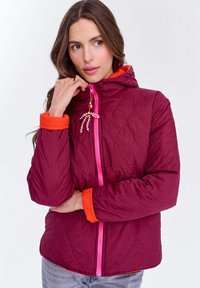 Kobieta w bordowej pikowanej kurtce z pomarańczowymi mankietami i podszewką kaptura, trzymająca zamek błyskawiczny z ozdobnym breloczkiem blisko swojej twarzy.