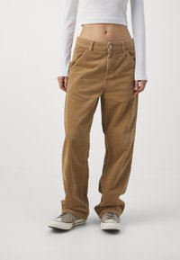 Pantalon en velours côtelé beige à jambes larges, doté d'une fermeture à boutons à l'avant et de poches latérales. Associé à un haut à manches longues blanc et des baskets.
