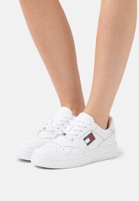 Bílé kožené tenisky s perforovanou špičkou, plochými tkaničkami a nášivkou s logem na boku v červené, bílé a modré barvě. Gumová podrážka, polstrovaný límec.