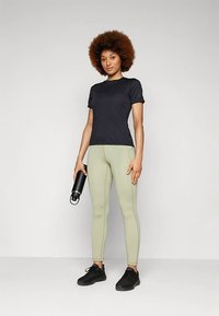 Νεαρή γυναίκα που στέκεται κρατώντας μια μαύρη μπουκάλι νερού, φορώντας ένα μαύρο μπλουζάκι, ανοιχτού πράσινου χρώματος κολάν και μαύρα αθλητικά παπούτσια, μπροστά από έναν απλό φόντο.