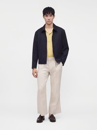 Jeune homme portant une veste noire, une chemise jaune boutonnée, un pantalon beige à jambes larges et des mocassins marron foncé, debout devant un fond uni.