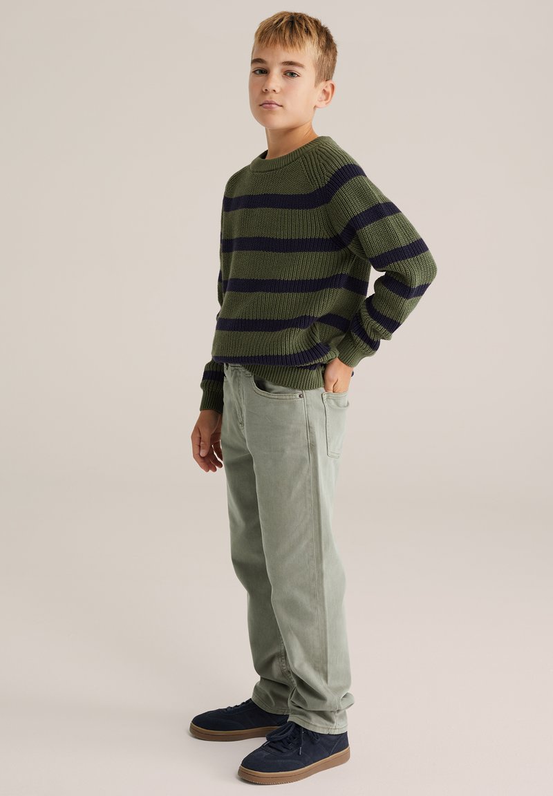 Zaļš un jūras debeszilas svītrains adīts džemperis ar ribotām piedurknēm, apvienots ar gaiši zaļām biksēm un ziliem zamšāda sporta apaviem. Stāvoša poza, neitrāls fons.