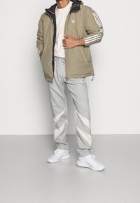 Beige vadderad jacka med marinblå foder och vita ränder, i kombination med grå joggingsbyxor med diagonala vita detaljer och vita sneakers.