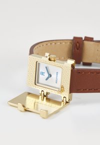 Gold square Tommy Hilfiger watch face flipped open on brown leather strap with beige stitching on white surface.