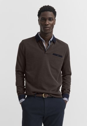 Uomo che indossa una polo marrone a maniche lunghe con colletto e polsini blu navy sopra una camicia a righe, abbinata a pantaloni blu navy e una cintura intrecciata.