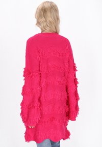 Cardigan tricotat supradimensionat fucsia, cu detalii de franjuri ondulați pe mâneci și margine, având o finisare texturată și o croială lejeră.
