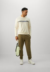 Lys beige sweatshirt med blå og grå stripe som har "LACOSTE," sammen med olivenfargede joggebukser, mens han holder en tenniskølle.