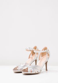 Srebrne sandały na wysokim obcasie z odkrytymi palcami, z plecionym wzorem z przodu, zapięciem na kostkę oraz smukłym, zwężającym się obcasem.