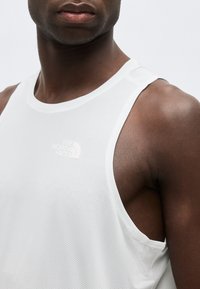 Κοντινό πλάνο ενός ατόμου που φοράει λευκό αμάνικο πλεκτό τοπ με το λογότυπο "The North Face" στο στήθος, με φόντο ένα απλό υπόβαθρο.