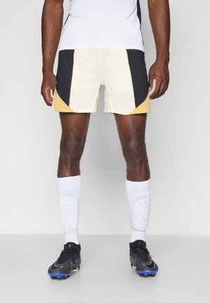 Atleet gekleed in witte, zwarte en gele sportshorts, witte kniehoge sokken en zwarte krammen, staand op een grijze vloer tegen een witte achtergrond.