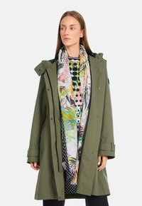 Olivgrüne wasserdichte Parka mit einem Reißverschluss und Druckknopfverschluss, verstellbare Kapuze und einem pinkfarbenen, grünen und schwarzen gemusterten Schal darunter.