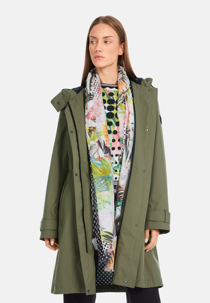 Olivgrüne wasserdichte Parka mit einem Reißverschluss und Druckknopfverschluss, verstellbare Kapuze und einem pinkfarbenen, grünen und schwarzen gemusterten Schal darunter.