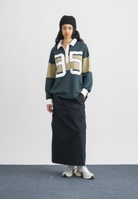 Zelena in bež dolg rokav polo majica z belim ovratnikom in velikim številom 95, kombinirana s črno krilom v stilu cargo in belimi supergami.