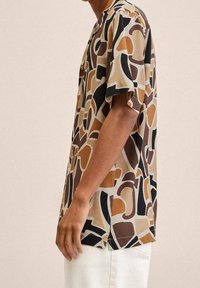 Mann, der ein kurzärmliges Hemd mit abstrakten braunen, schwarzen und beigen Formen trägt, kombiniert mit weißen Hosen, seitlich stehend.