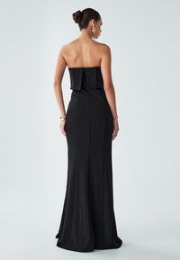 Melnas strapless kleitas ar pieguļošu augšu un izplestu apakšdaļu, ar aizmugures rāvējslēdzēju un smalku drapējumu, kas nodrošina gludu siluetu.