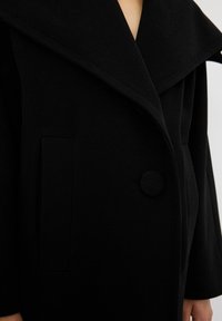 Cappotto di lana nera con un ampio colletto ripiegato, chiusura a un bottone e tasca laterale. Texture morbida e design strutturato.