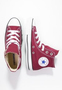 Baskets montantes en toile bordeaux avec des semelles en caoutchouc blanches, arborant un patch circulaire bleu et des lacets blancs, présentées en vue de dessus.