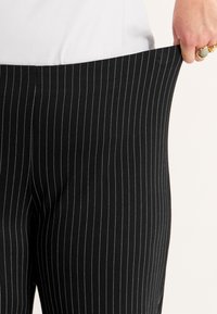 Persoon trekt de tailleband van losse zwarte pinstripe pants, draagt een witte top en gouden ringen aan de rechterhand.