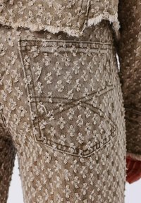 Los jeans de mezclilla beige presentan un patrón texturizado con pequeños diseños elevados. El bolsillo trasero es visible, junto con el detalle de dobladillo deshilachado.