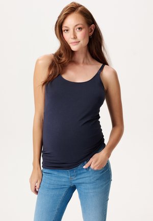 Schwangere Frau mit langen braunen Haaren, die ein marineblaues Tanktop und blaue Jeans trägt, mit einer Hand in der Tasche stehend vor hellem Hintergrund.