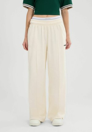Osoba ubrana w luźne kremowe spodnie dresowe z elastycznym paskiem w talii i małym czarnym logo, w połączeniu z białymi trampkami i ciemnym krótko skrojonym topem.