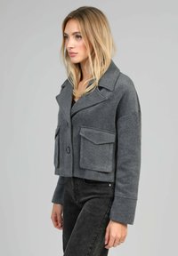 Veste grise courte avec de grandes poches avant, un col cranté et une fermeture à bouton, fabriquée dans un tissu doux et texturé.