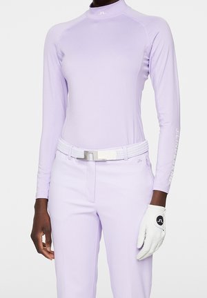 Persona vistiendo un conjunto ajustado de camiseta de manga larga lavanda y pantalones, con un cinturón trenzado blanco y un guante de golf blanco en la mano izquierda, sobre un fondo liso.