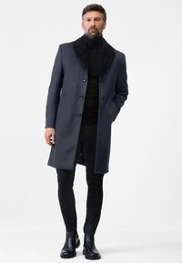 Heren lange jas van donkergrijze stof, met een kraag van zwart nepbont, voorzijde met knopen en zijzakken, gecombineerd met zwarte broek en enkellaarsjes.