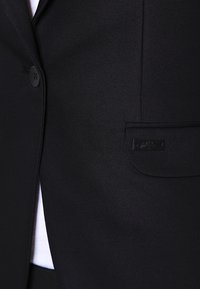 Blazer nero con una texture liscia, chiusura con un solo bottone e una piccola etichetta rettangolare sul taschino sinistro.