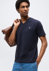 Άνδρας με μπλε σκούρο T-shirt με V-λαιμόκοψη και λευκό λογότυπο, μπλε τζιν, κρατώντας καφέ δερμάτινο μπουφάν στον ώμο, κοιτάζοντας δεξιά σε λευκό φόντο.