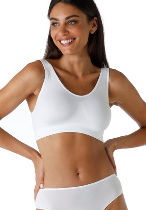 Donna sorridente con lunghi capelli scuri che indossa un reggiseno sportivo bianco senza cuciture e mutandine bianche abbinate su uno sfondo bianco.