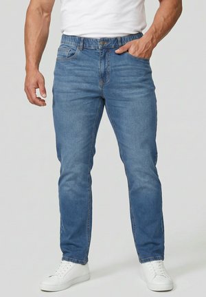 Mann trägt blaue Jeans und weiße Turnschuhe, steht mit einer Hand in der Tasche und der anderen neben dem Körper.