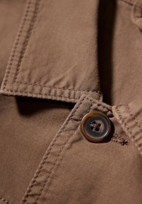 Brunt tyg med en texturerad yta, med en sydd krage och en stor rund knapp nära sömmen som framhäver detaljerat hantverk.